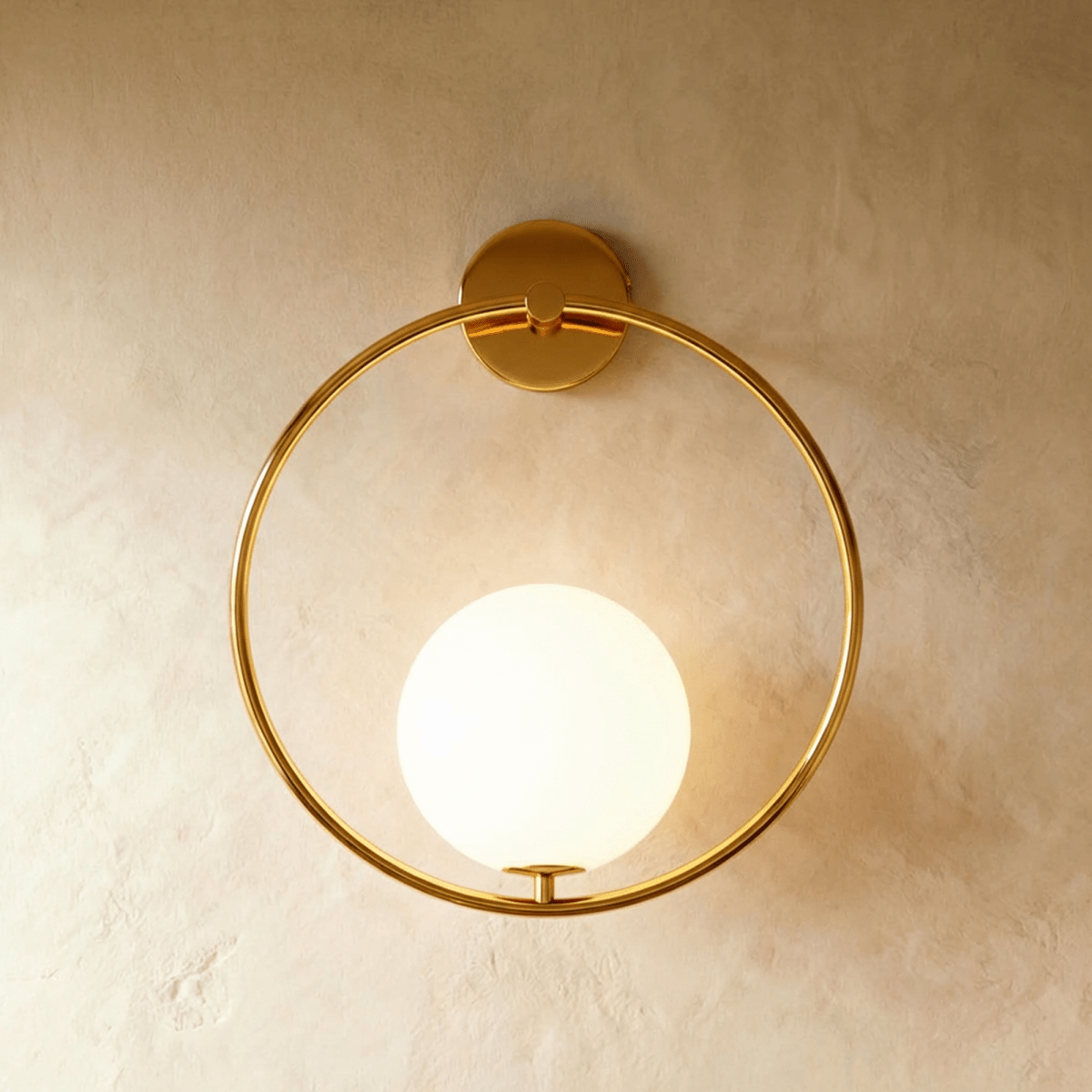 Clérachic wall lamp in gold, a full hoop hanging from a round wall plate with the lit sphere seated low inside it on a short stem.
