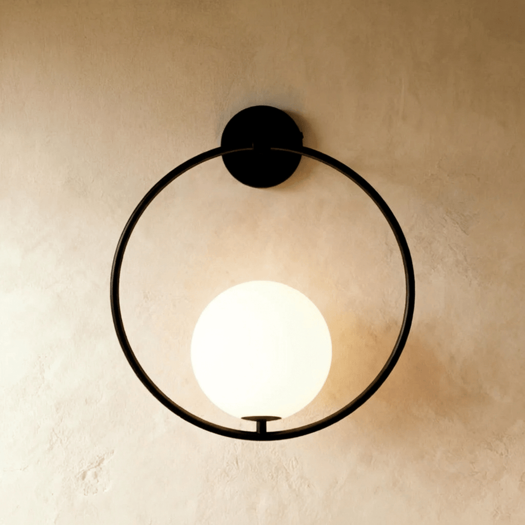 Clérachic wall lamp in matte black, the same hoop and low-seated lit sphere against a warm plaster wall.