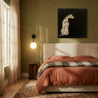Clérachic wall lamp in black, lit beside a bed on an olive green wall above a wavy-edged wooden side table.