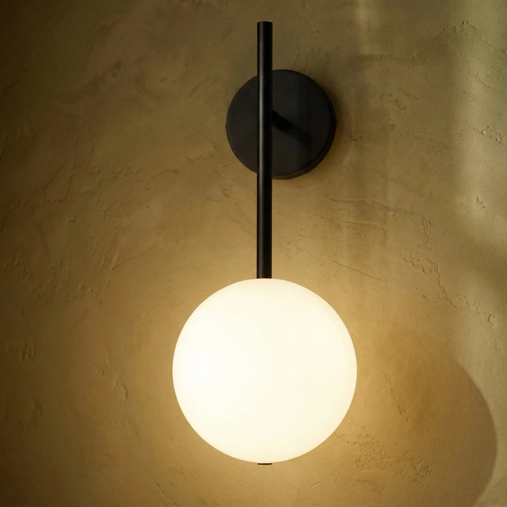 Clérachic wall lamp detail in black, the straight rod and warmly lit sphere seen close against textured plaster.