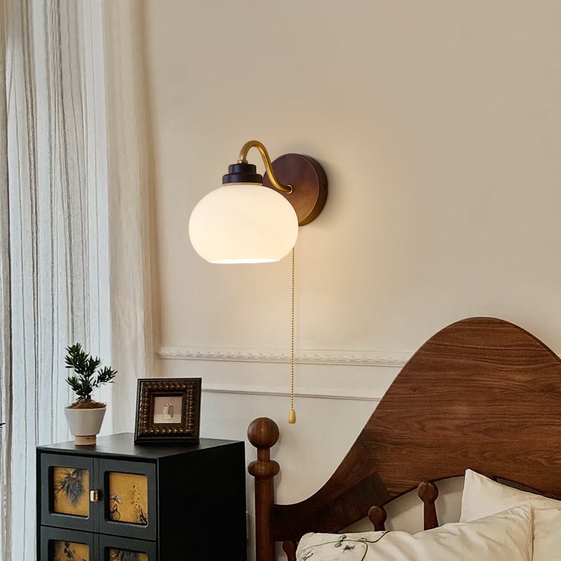 A Clérachic wall light with a smooth rounded glass shade on a curved gold arm, over a carved wooden bed and a black cabinet.
