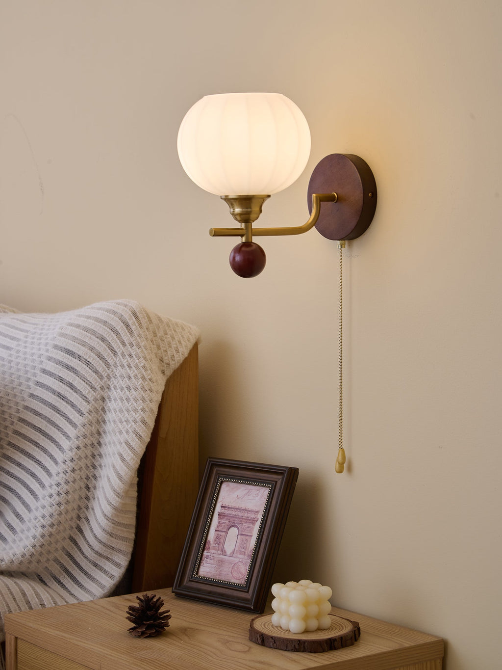 Clérachic wall light with a ribbed globe, lit gently, above a pale wood bedside table with a framed print and a small candle.