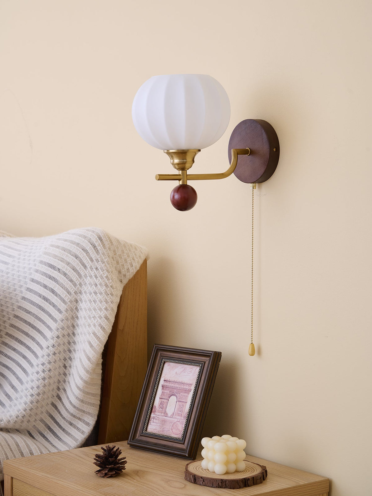 Unlit ribbed white glass globe of this Clérachic wall light, above a bedside table with a framed print and a sculpted candle.