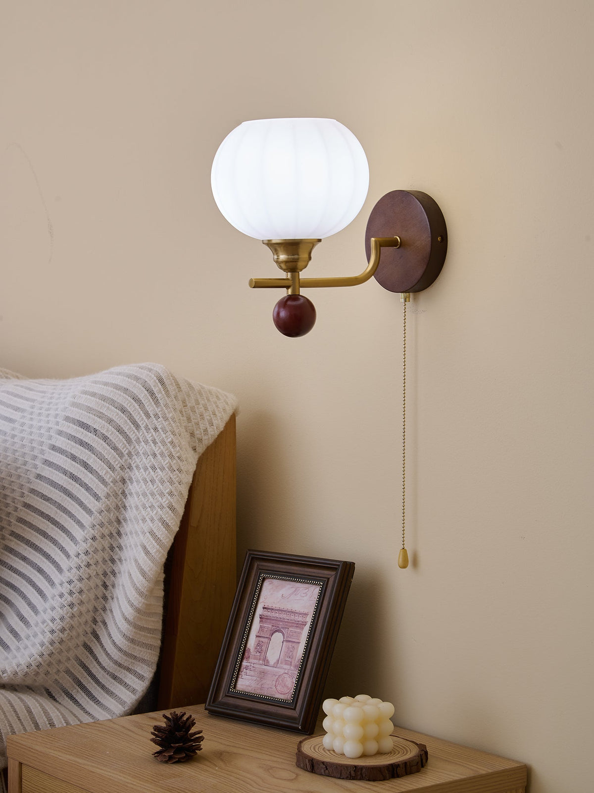Clérachic wall light with a bright white ribbed globe, seen in daylight above a bedside table with a framed print and pine cone.