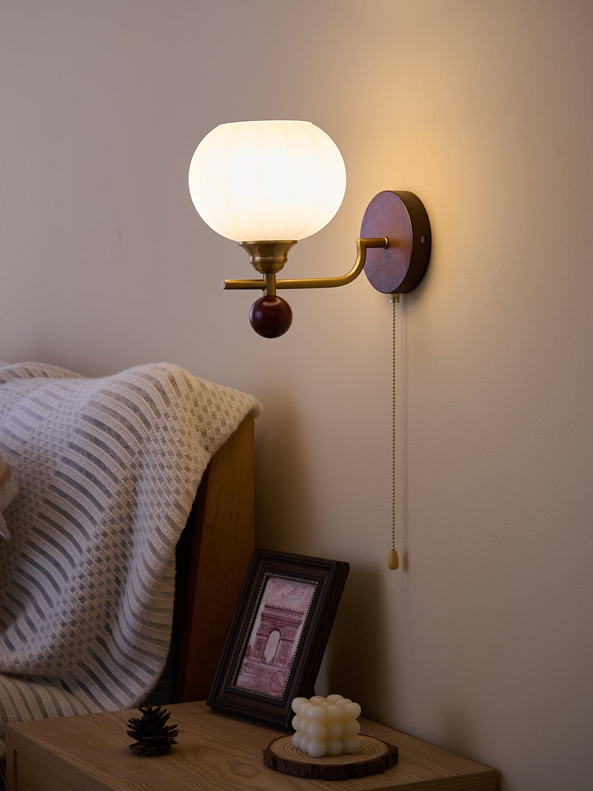 Ribbed globe Clérachic wall light glowing warm against a dusky taupe wall, above a bedside table with a framed print.