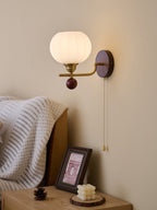 A Clérachic wall light with a ribbed frosted globe and pull chain, hung beside a bed with a white knitted blanket.