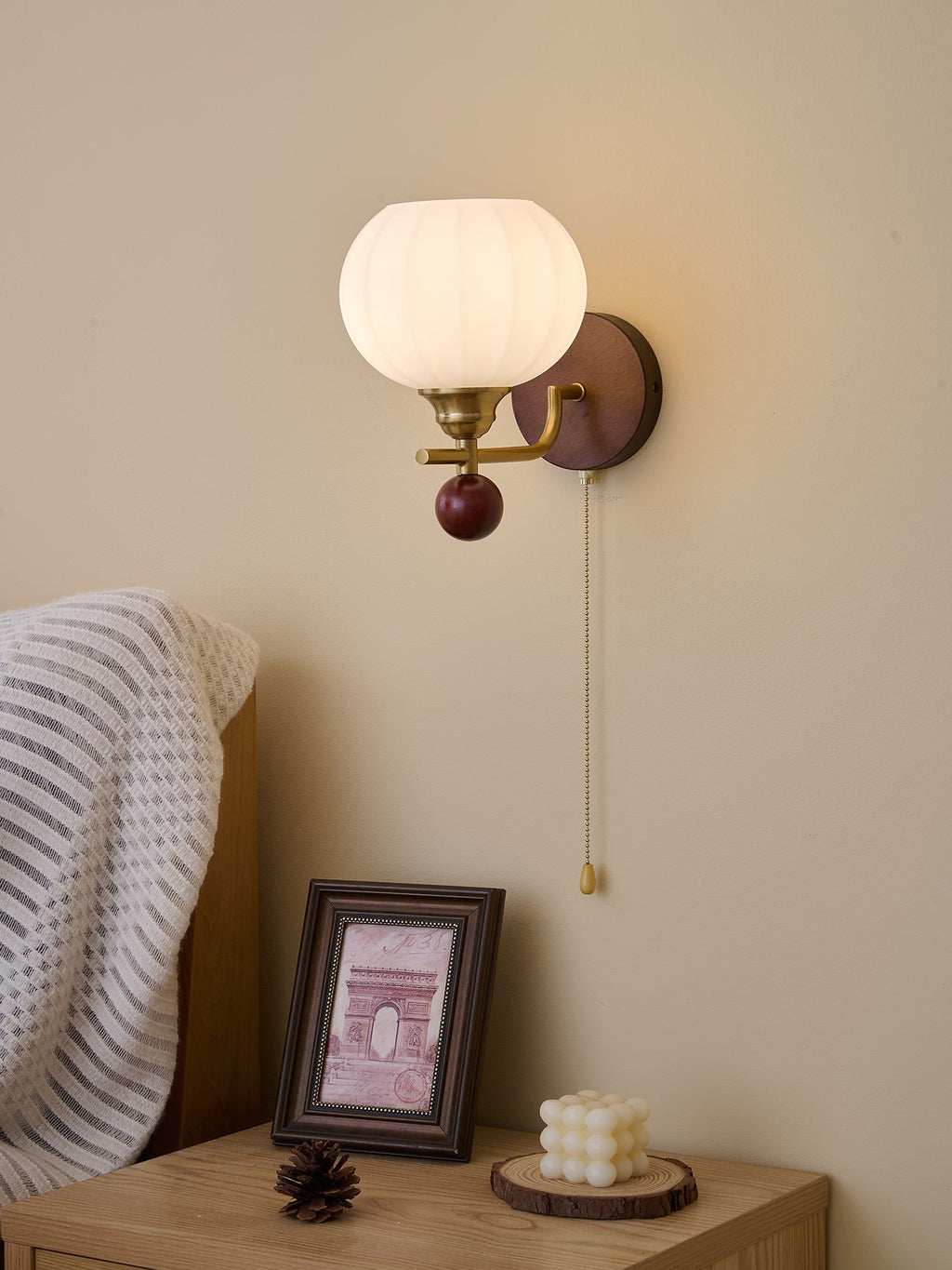 Clérachic wall light lit warm in a softly shaded bedroom, with the chain hanging down by a framed print and a pine cone.