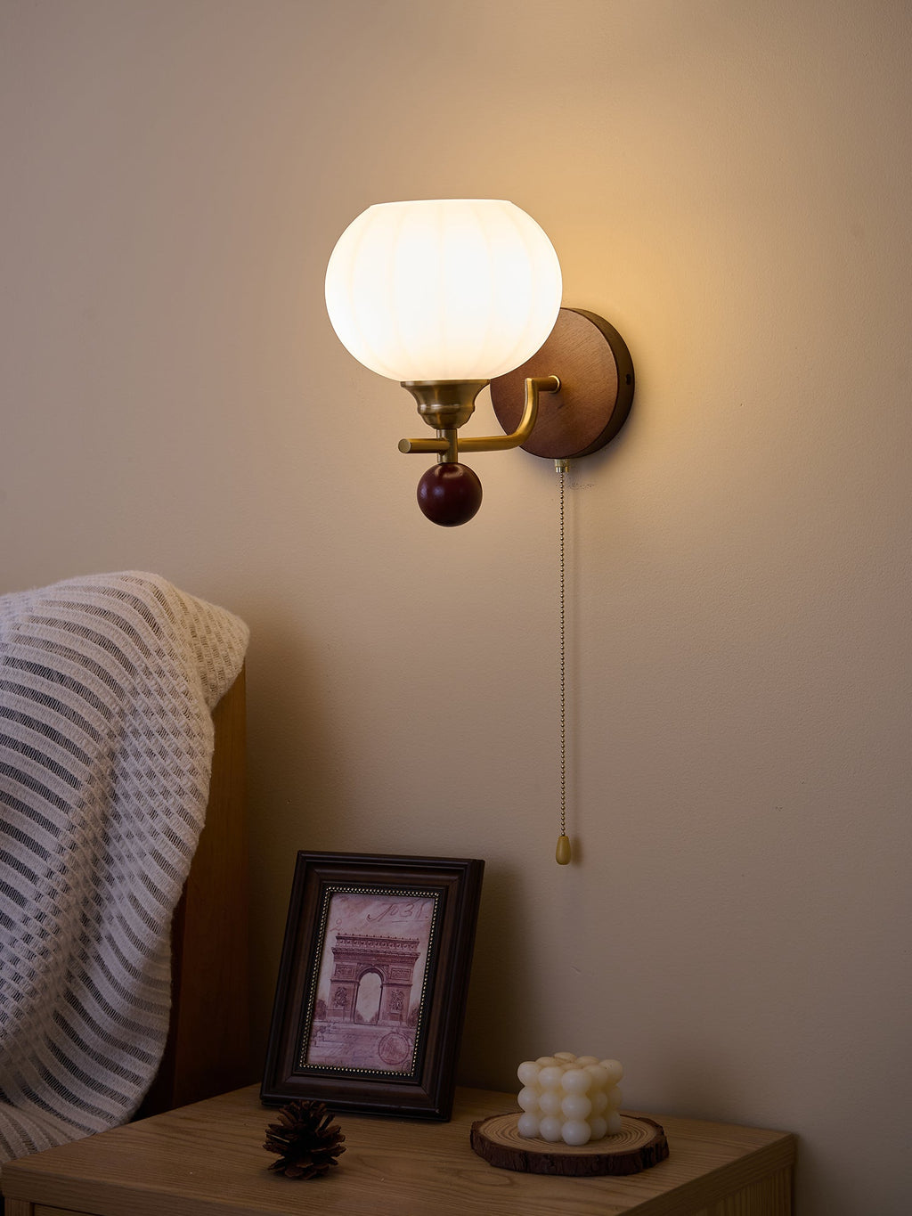Ribbed Clérachic wall light shining brightly in a darker room, with a glow around the globe and a bedside table below.