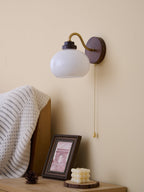 A Clérachic wall light with a smooth, round white glass shade on a curved gold arm, above a bedside table and framed print.