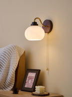 Smooth glass shade of this Clérachic wall light lit on a pale wall, with a curved gold arm and a pull chain beside a framed print.