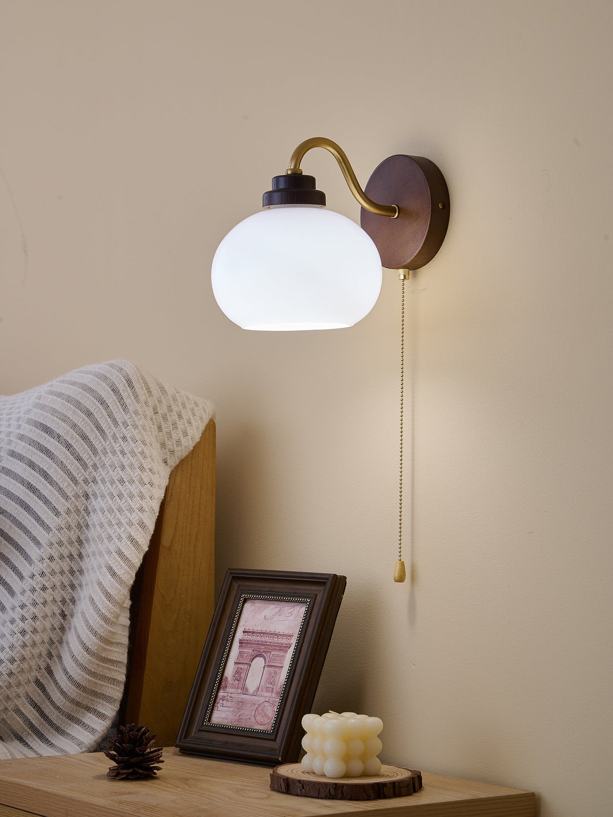 Brightly lit Clérachic wall light with a smooth white glass shade and a curved gold arm, above a bedside table.