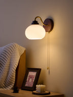 A Clérachic wall light with a smooth glass shade casts a warm glow on a dusky wall, above a bedside table and a framed print.