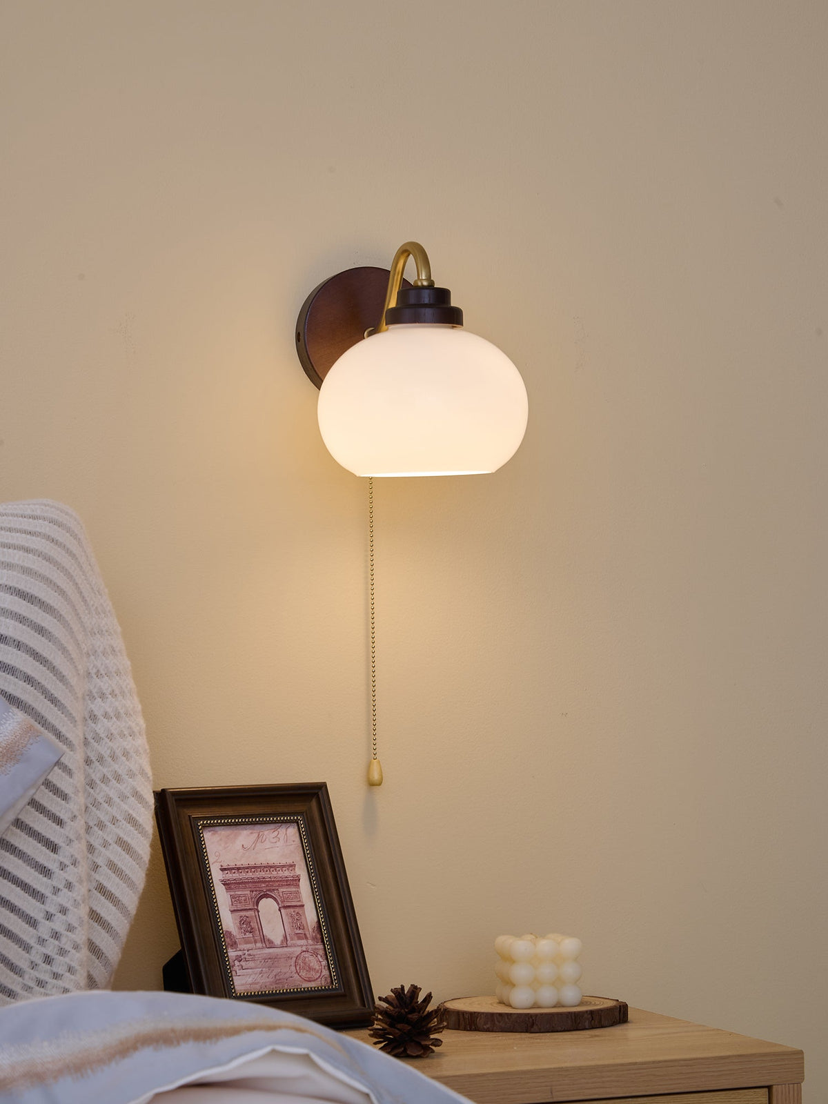 Clérachic wall light with the curved arm on the left and a smooth glass shade, lit warm above a framed print and a candle.