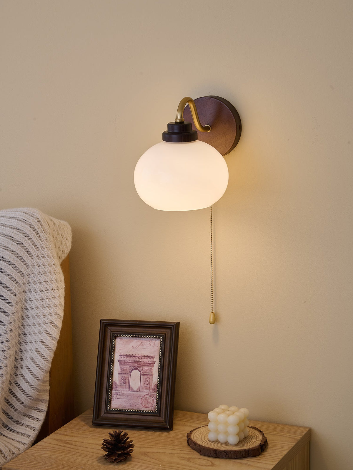 Smooth, rounded glass shade on a Clérachic wall light, lit softly beside a bed with a knitted throw and a framed print.