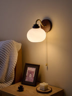 Clérachic wall light with a smooth glass shade glowing against a muted wall, with a framed print and a candle on the table below.