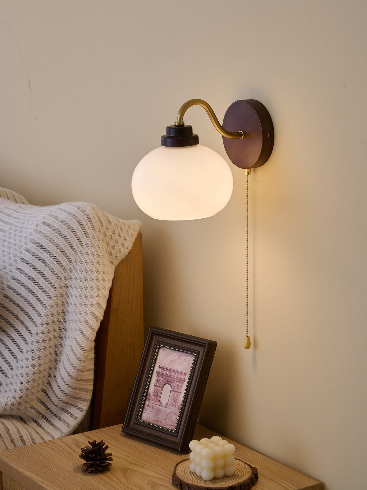 A Clérachic wall light with a smooth round shade and a dark top cap, hung close to a bed with a white knitted throw.
