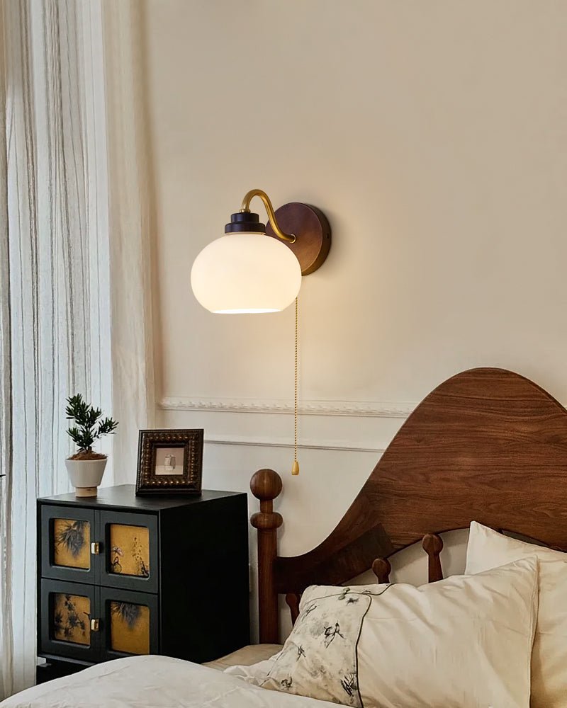 Clérachic wall light with a smooth glass shade on a curved arm, beside sheer curtains and a carved wooden bed with a black cabinet.