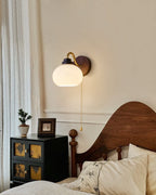 Clérachic wall light with a smooth glass shade on a curved arm, beside sheer curtains and a carved wooden bed with a black cabinet.