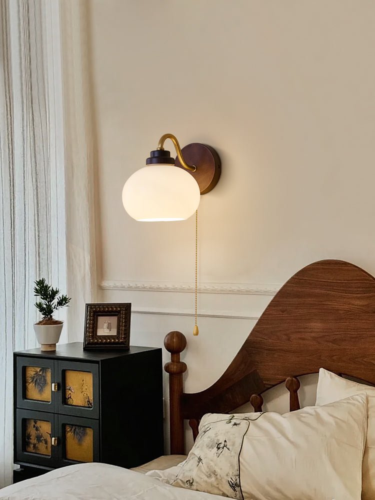 A Clérachic wall light with a smooth glass shade above a black bedside cabinet with gold panels, with sheer curtains to the left.