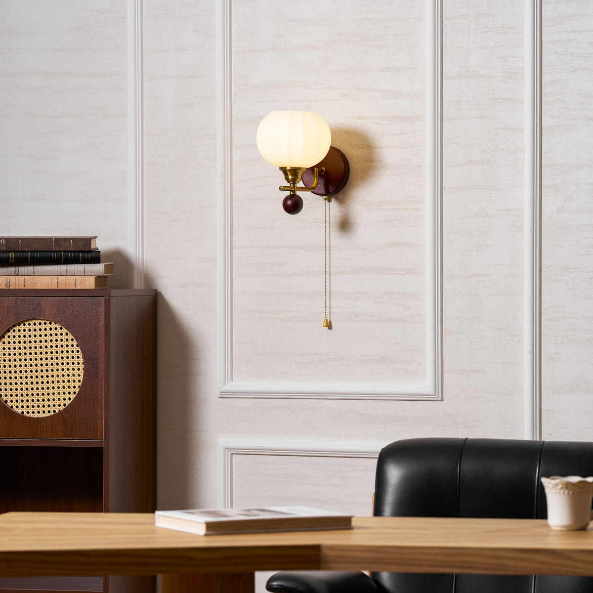 Clérachic wall light lit on a white panelled wall, above a rattan-fronted dark wood cabinet and a leather chair at a desk.
