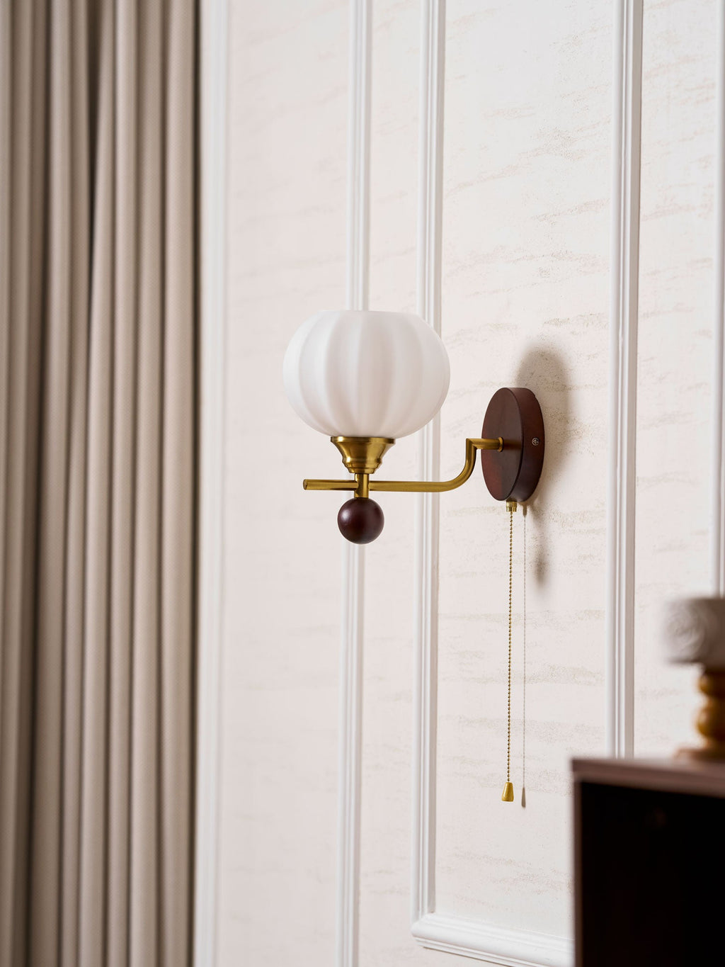 Ribbed globe Clérachic wall light beside a taupe curtain on a white panelled wall, with a small dark cabinet at the edge.