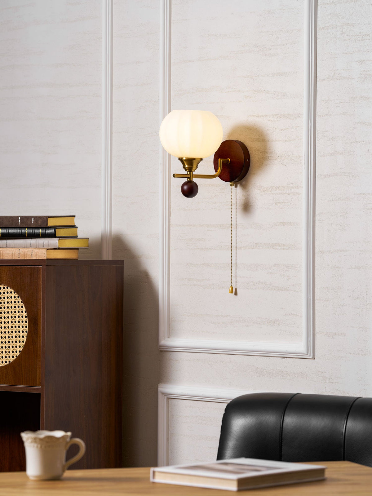 A Clérachic wall light high on a panelled wall, above a rattan-panel cabinet, a teacup, an open book and a leather chair.