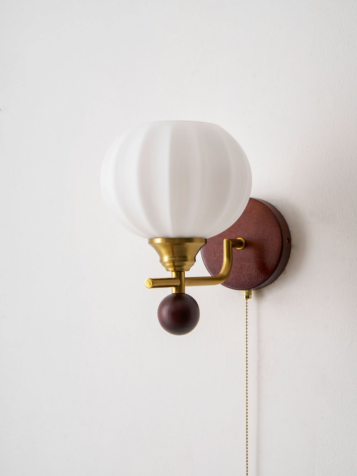 Close view of the ribbed white glass globe, gold arm and walnut-toned ball on this Clérachic wall light, against a plain white wall.