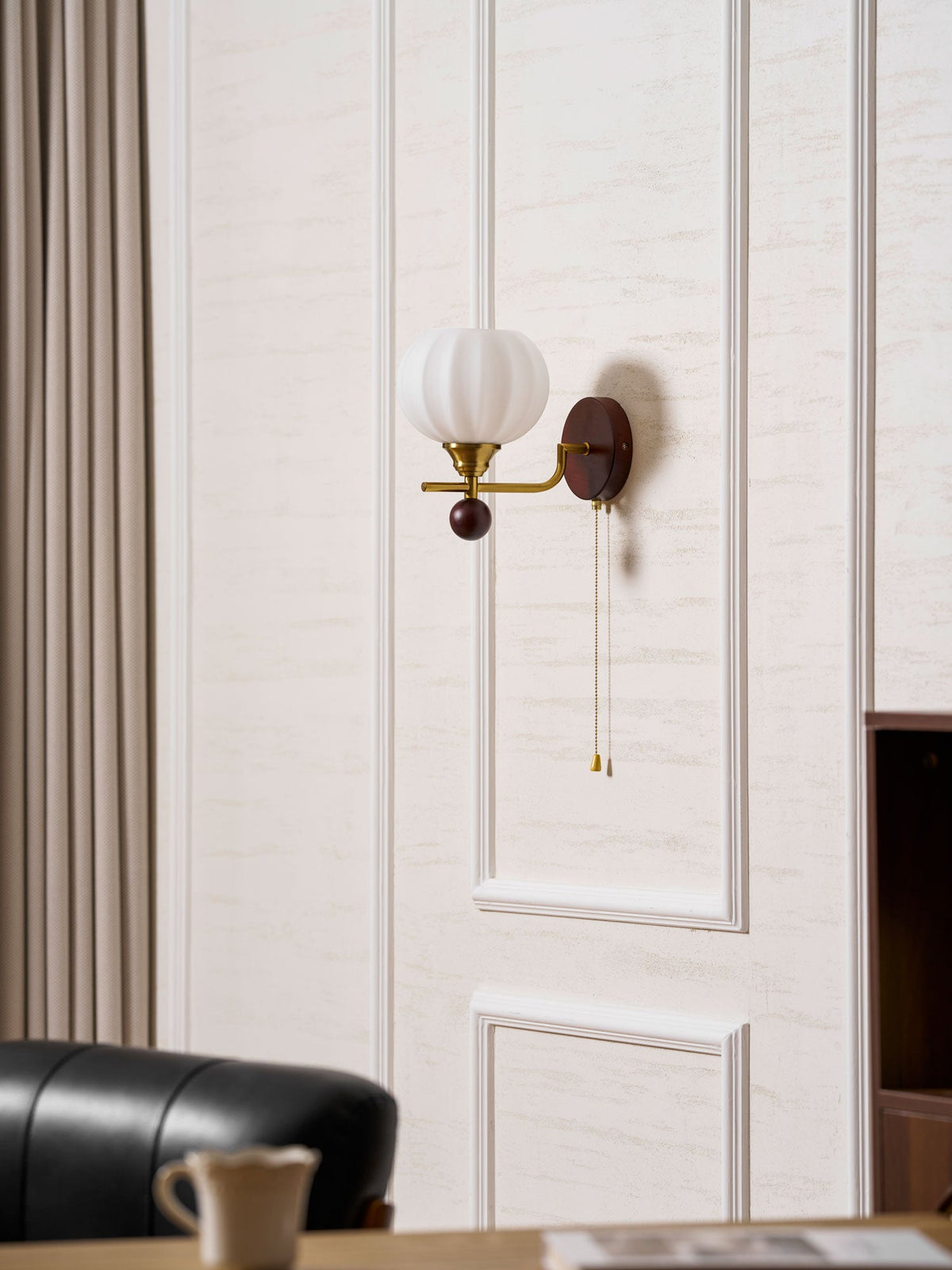 A Clérachic wall light on white wall panels, with a taupe curtain at the side and a leather chair and a teacup below.
