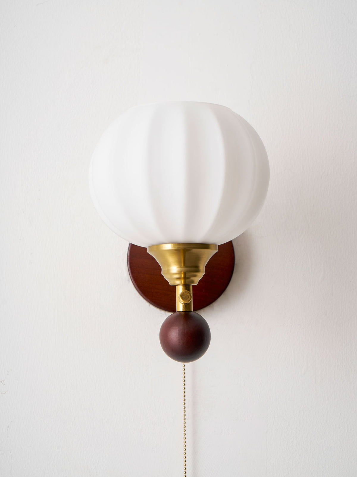 Front view of the ribbed glass globe on this Clérachic wall light, with its gold fitting, walnut-toned ball and pull chain.