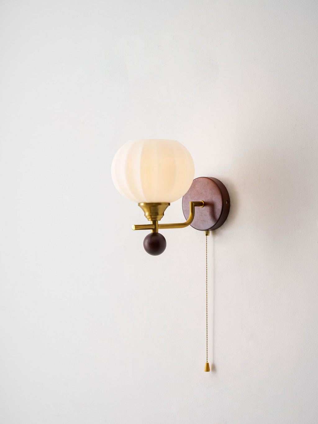 Side-on view of a lit Clérachic wall light with a ribbed globe, a gold arm and a white pull cord with a gold tip, on a pale wall.