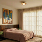 Clérachic wood-grain disc ceiling light with a globe glowing above a bed with a rose cover, beside tall sheer-curtained windows.