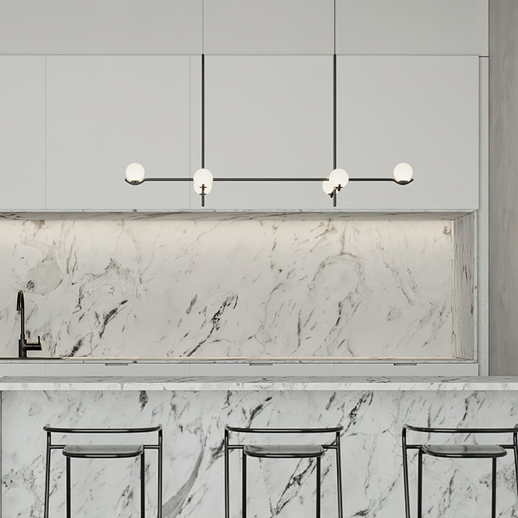 A Clérachic linear chandelier with frosted globes hangs in a white kitchen, above a marble island with black stools.
