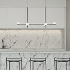 A Clérachic linear chandelier with frosted globes hangs in a white kitchen, above a marble island with black stools.