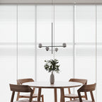 A Clérachic chandelier with four rounded shades hangs before tall white blinds above a round table and an olive branch.