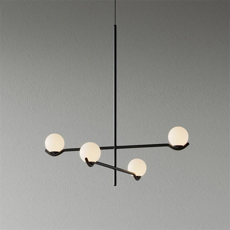 This Clérachic chandelier in black, with four frosted globes on crossing arms, on a plain grey wall.