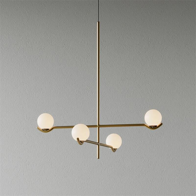 This Clérachic chandelier in gold, with four frosted globes on crossing arms, on a plain grey wall.