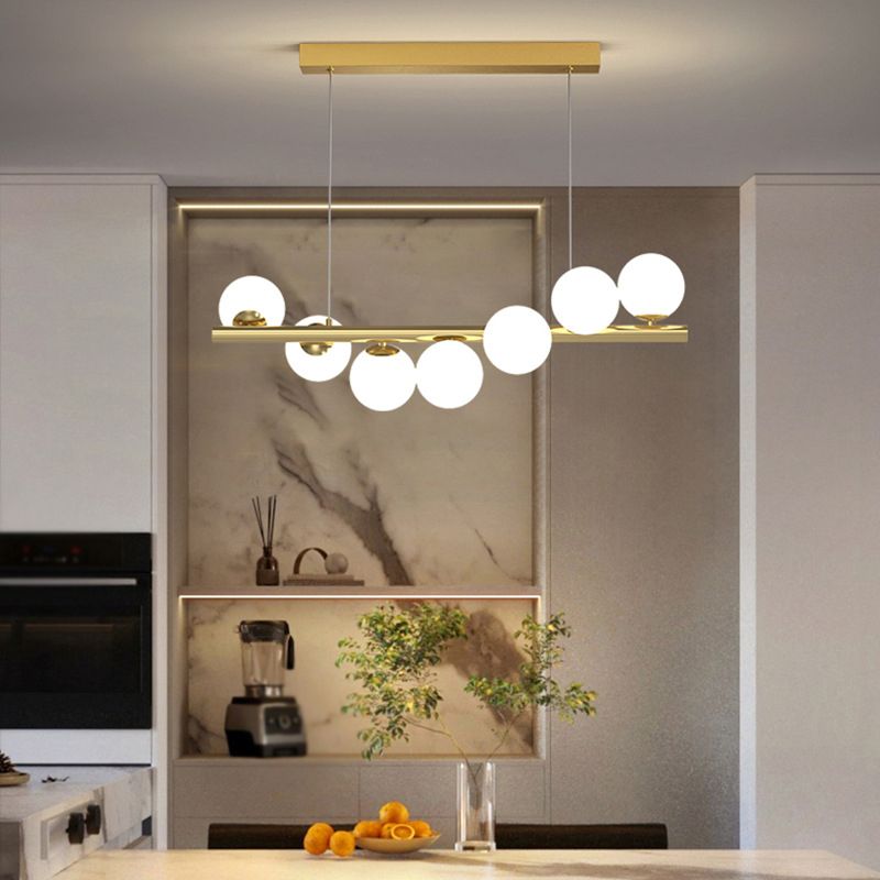 The gold Clérachic Frosted Globe Linear Pendant Light hangs over a kitchen island with a marble backsplash and fresh greenery.