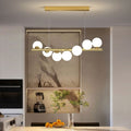 The gold Clérachic Frosted Globe Linear Pendant Light hangs over a kitchen island with a marble backsplash and fresh greenery.