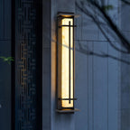 This Clérachic wall light glows warm in its dark frame against a grey stone wall at dusk, beside a carved lattice panel.