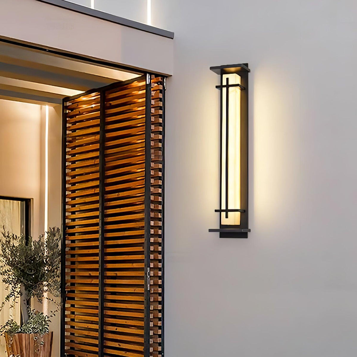 A Clérachic wall light lit on a pale rendered wall beside a doorway with timber slats and a potted olive tree.
