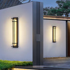 Two Clérachic wall lights glowing on white rendered walls beside a dark column, with clipped hedging below at dusk.