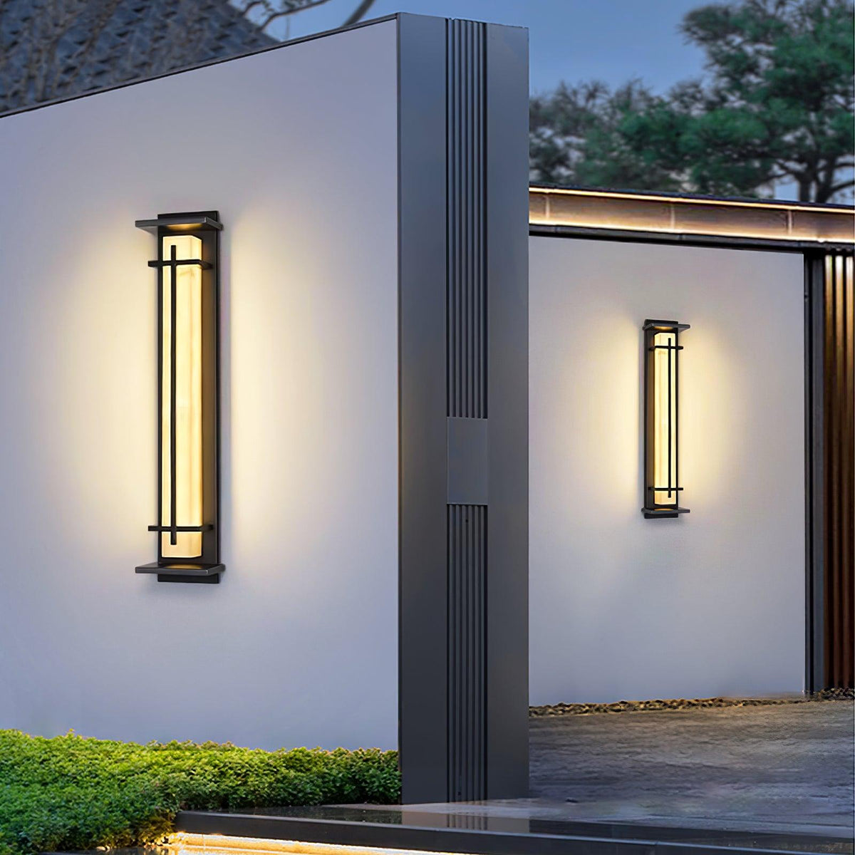 Two Clérachic wall lights glowing on white rendered walls beside a dark column, with clipped hedging below at dusk.