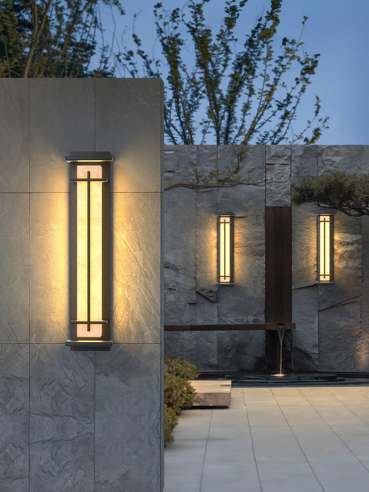 Dark-framed wall light with a glowing alabaster-style shade on a slate wall, a pair of smaller lights beyond; from Clérachic.