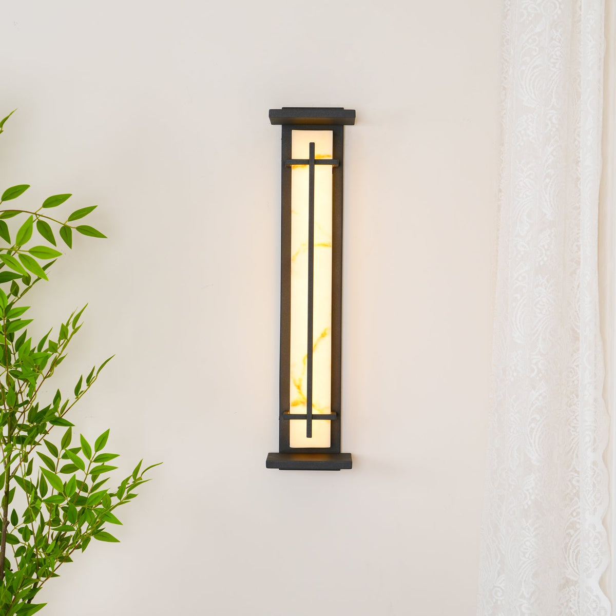 This Clérachic wall light on a cream wall, switched off, with its dark frame and a green plant to one side.