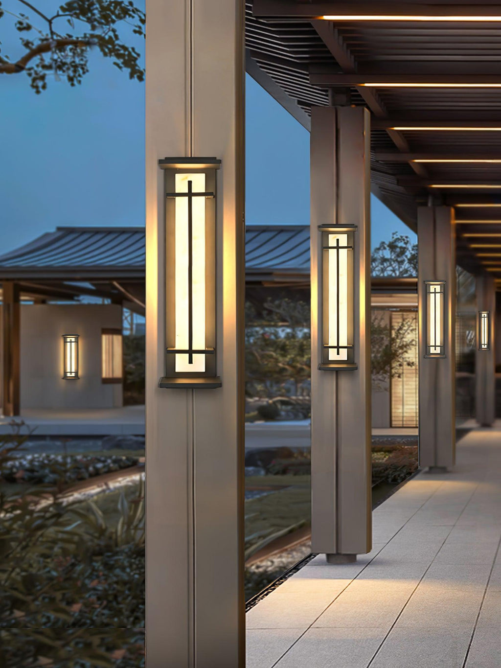 Lit Clérachic wall lights on the columns of a covered walkway, glowing warm toward a stone path and a garden at dusk.
