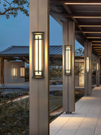 Lit Clérachic wall lights on the columns of a covered walkway, glowing warm toward a stone path and a garden at dusk.