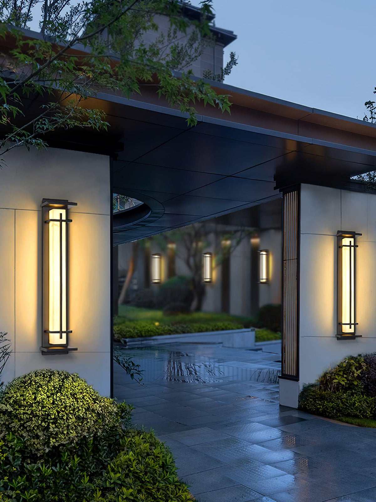 A Clérachic wall light on a pale pillar by a garden entrance, with a second light opposite and shrubs and wet paving below.