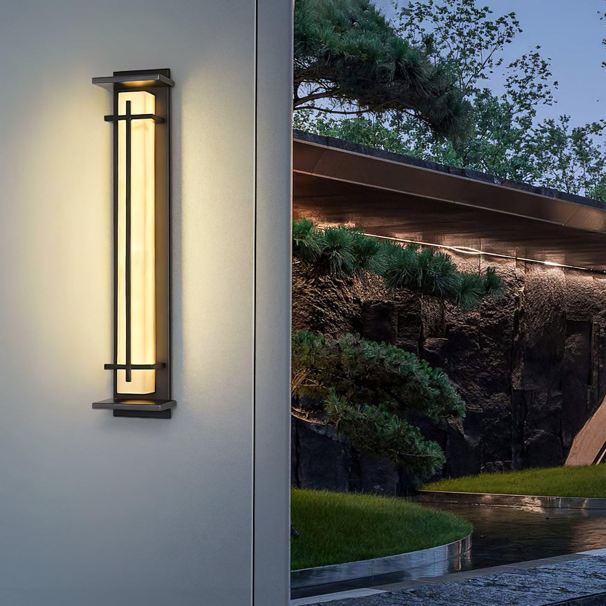 Large warm-glowing wall light on a grey wall, with a pine tree and a low roofline in the background; a Clérachic design.