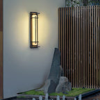 This Clérachic wall light casts a warm halo on a grey wall beside a timber fence and a standing stone with low planting.