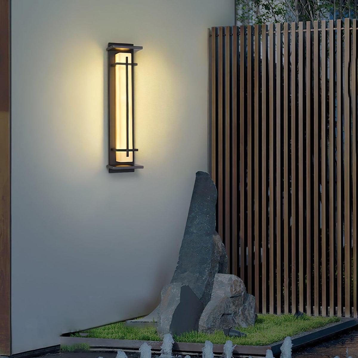 This Clérachic wall light casts a warm halo on a grey wall beside a timber fence and a standing stone with low planting.
